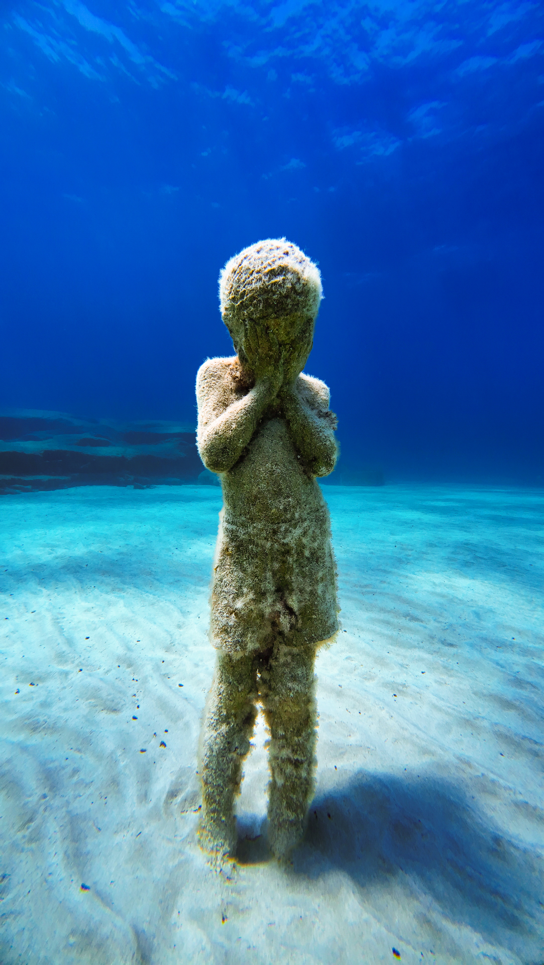 A submerged child sculpture at MUSAN, colonised by fine marine growth, standing on the pale sandy seabed.