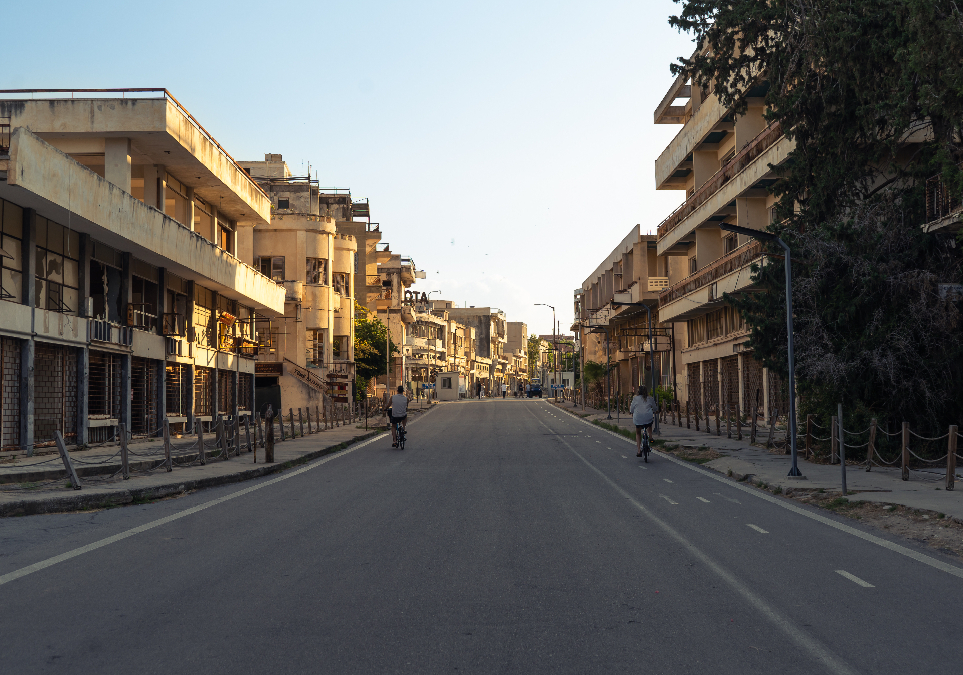 A wide, empty avenue in Varosha at golden hour, lined with crumbling mid-century buildings as two cyclists ride into the light.