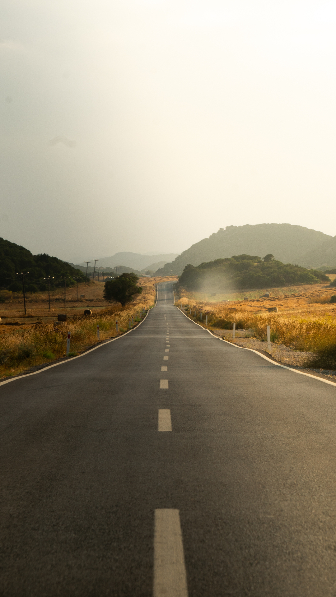 An empty tarmac road running straight through open fields toward distant hills at sunset.