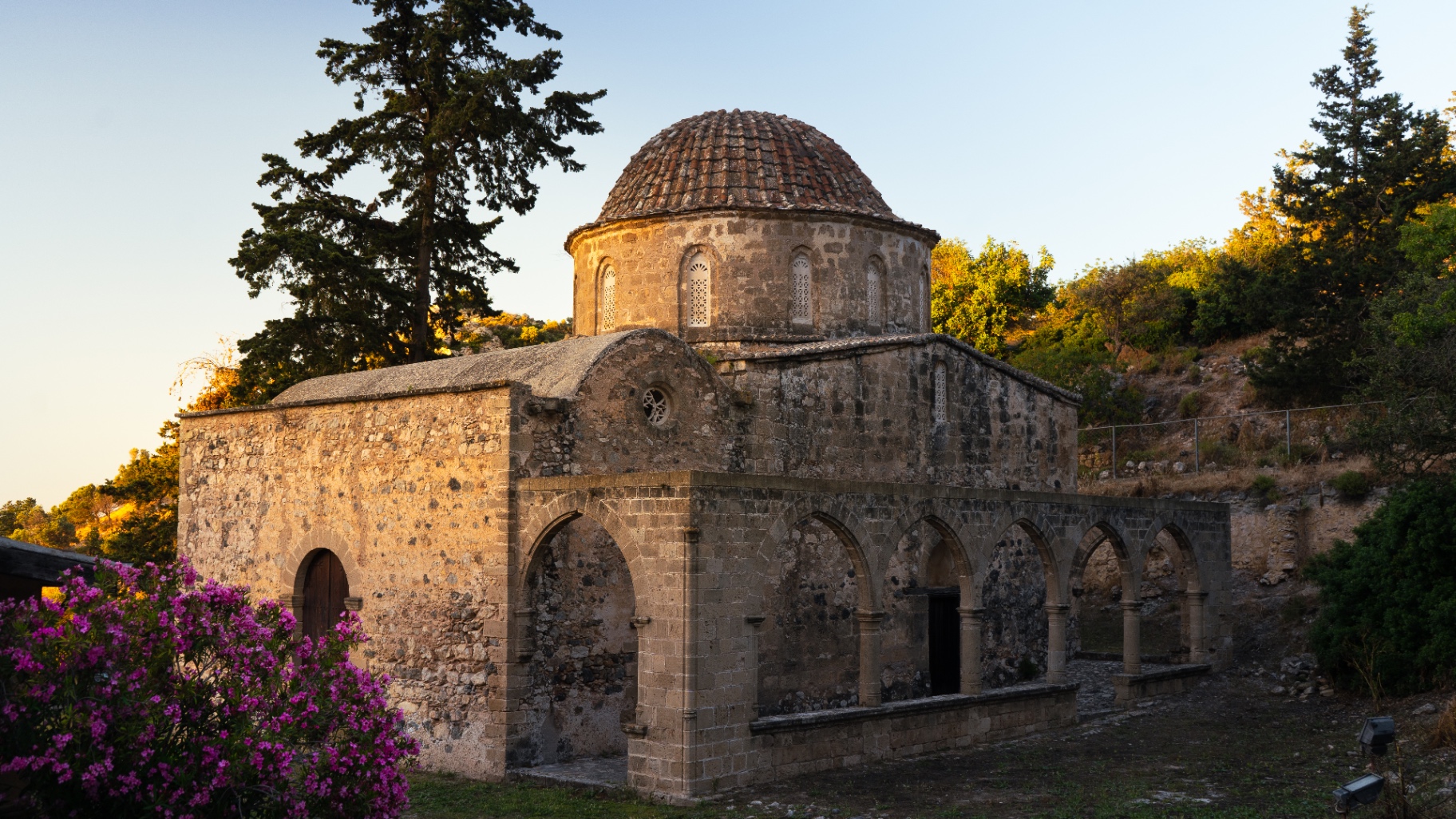 A small Byzantine stone chapel with a tiled dome glowing in evening light, framed by cypress trees and pink oleander.