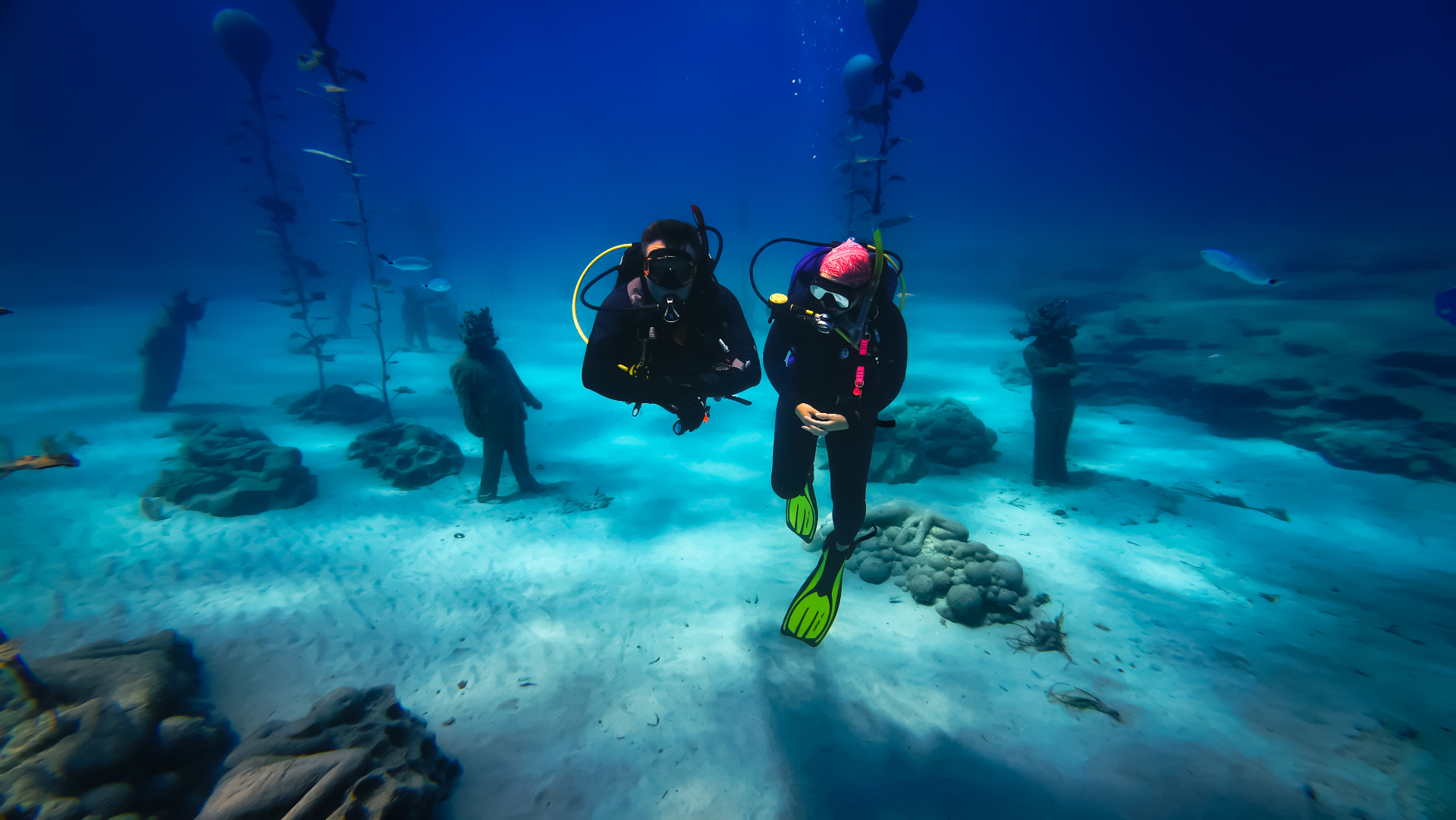 Two divers finning slowly between the sculptures and trees of the MUSAN installation.