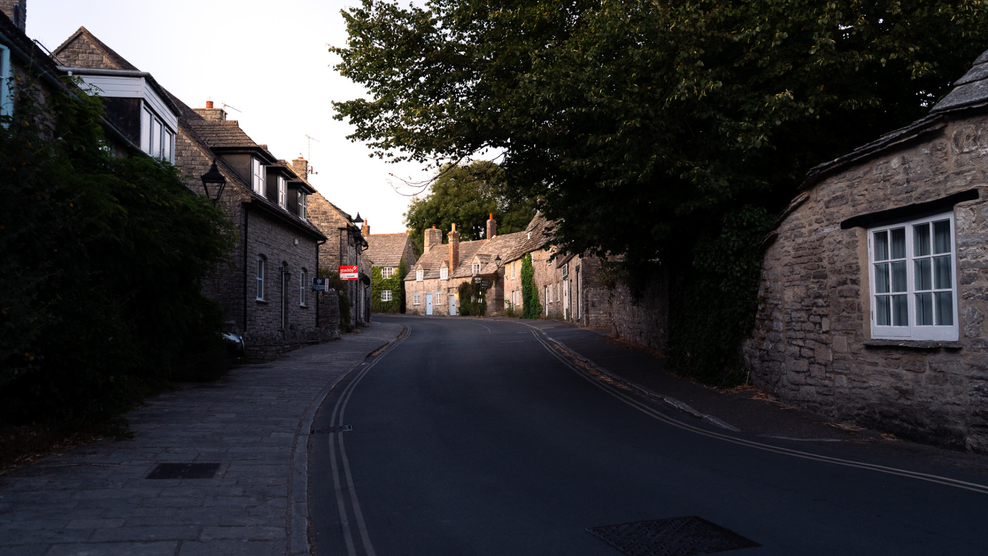 An empty road curving between stone cottages in a Dorset village at dusk.