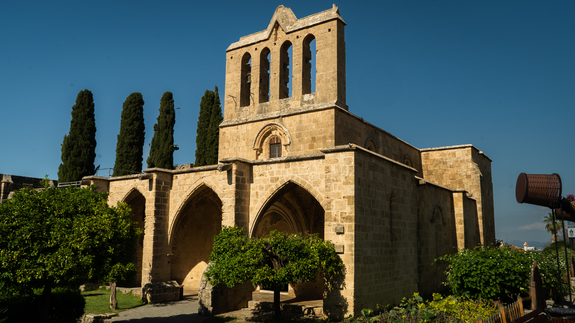 The stone bell tower and Gothic arches of Bellapais Abbey rising above cypress trees under a clear blue sky.